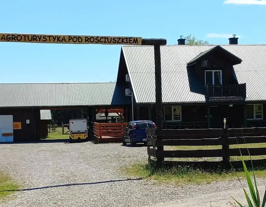Brama z napisem Agroturystyka Pod Rościuszkiem, za nią drewniany domek noclegowy w słońcu