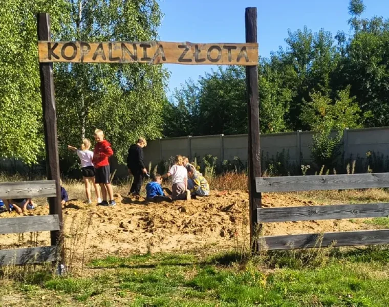 Dzieci kopią w piasku pod drewnianą bramą z napisem Kopalnia złota